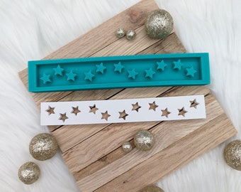 May include: A teal silicone mold with 15 star-shaped cavities and a white plastic template with 15 star-shaped cutouts. The mold and template are on a wooden surface with gold glitter ornaments.