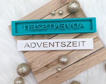 May include: A teal blue silicone mold with the word "ADVENTSZET" spelled backwards. A white wooden sign with the word "ADVENTSZET" is below the mold. There are several gold glitter ornaments on a wooden surface.