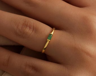 May include: A gold ring with a rectangular emerald gemstone set in the center. The ring is thin and delicate, with a polished finish. The emerald is a vibrant green color, and the gold band is smooth and shiny.