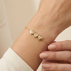 Könnte beinhalten: Ein goldfarbenes Armband mit drei herzförmigen Anhängern. Jeder Anhänger hat einen Buchstaben eingraviert. Das Armband wird am Handgelenk getragen, die Kette ist zart und dünn.