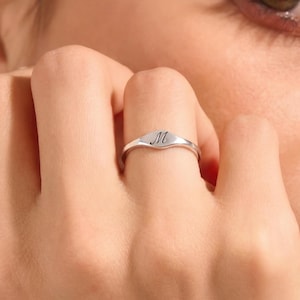 May include: A silver-coloured metal ring featuring a smooth, polished band that widens slightly at the centre. The top surface of the ring is engraved with the uppercase letter M in a classic, elegant script font, centred on the metal face.