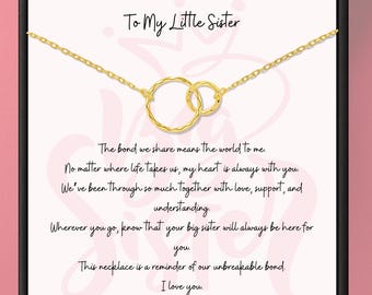 Könnte beinhalten: Eine goldfarbene Halskette mit zwei ineinandergreifenden Kreisen, präsentiert auf einer Karte mit dem Text "To My Little Sister". Die Karte enthält eine herzliche Botschaft über die Schwesternschaft. Die Halskette ist ein liebevolles Geschenk.