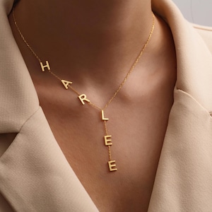 May include: A gold-colored necklace spelling out the name "HARLEE" in individual block letters. The letters are connected by a delicate chain, with the name cascading down the neckline. The necklace is worn against a neutral-toned blazer.