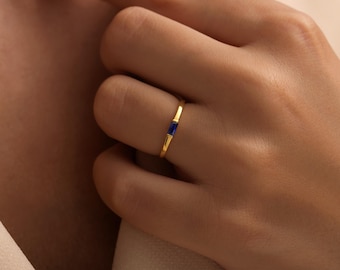 May include: A gold ring with a rectangular, dark blue gemstone. The ring is thin and delicate, with a simple band. The gemstone is set horizontally in the center of the ring. The ring is on a finger.