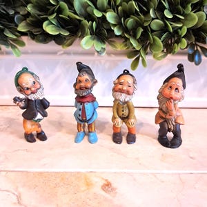 May include: Four small, vintage gnome figurines with white beards and colorful outfits. The gnomes are wearing black, blue, tan, and brown clothing. They are standing on a marble surface, with a green leafy garland in the background.