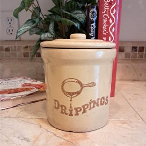 May include: A beige ceramic crock with a lid, featuring a brown illustration of a pan and the word "DRIPPINGS." The crock is on a tiled surface, with cookbooks and greenery in the background. A vintage kitchenware item.