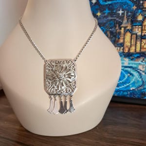 May include: Silver-toned necklace with a rectangular pendant featuring an ornate, openwork design. The pendant has a central starburst pattern and three dangling, teardrop-shaped charms. The necklace is displayed on a mannequin.