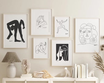 May include: A collection of framed art prints featuring minimalist line drawings in black and white. The artwork includes abstract figures and faces, displayed above a white cabinet with a lamp, decorative objects, and a vase with dried plants. A bed with a gray comforter is in the foreground.