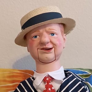 May include: A vintage ventriloquist dummy doll with a straw hat, wearing a navy blue and white striped suit, a white collared shirt, and a red polka dot tie. The doll has a painted face and is seated on a floral patterned surface.