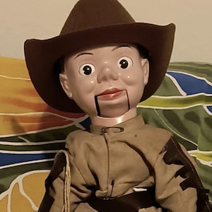 May include: A vintage ventriloquist doll with a painted face, wearing a brown cowboy hat and a tan outfit with fringed accents. The doll has a string-operated mouth and is standing on a white base.