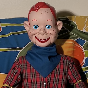 May include: A ventriloquist dummy with red hair, blue eyes, and freckles. The doll wears a red and black plaid long-sleeve shirt, a solid blue neckerchief, a red belt with a circular buckle, and solid blue trousers with gold-colored shoes.