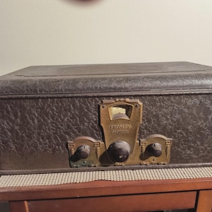 May include: A vintage Crosley Showbox radio in a brown textured case. The radio has brass-colored knobs and a dial. The case is rectangular with a slightly rounded top. The radio sits on a wooden table.