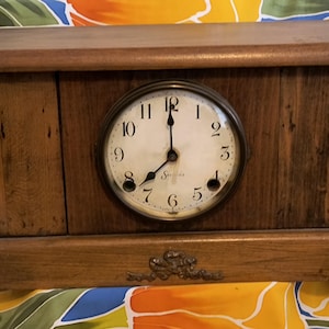 May include: A wooden mantle clock with a round clock face. The clock face has black numerals and hands, and the clock is housed in a rectangular wooden case. The clock has decorative accents on the bottom.