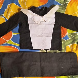 May include: A black and white costume set, including a long-sleeved top with a white bib and a pair of matching black pants. The outfit is laid out on a colorful floral patterned fabric.