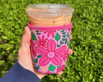 May include: A pink floral coffee cup sleeve with a clear plastic lid, holding an iced coffee. The sleeve features a repeating pattern of smiling flowers, green leaves, and yellow stars. The rim of the sleeve is a darker pink.