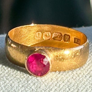 May include: A gold ring with a large, round, faceted ruby gemstone. The ring has a wide band and visible hallmarks. The ruby is a vibrant red color, and the gold has a slightly textured appearance.