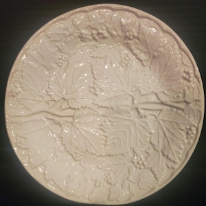 May include: A round, off-white ceramic serving platter with a raised grape and leaf design. The platter features detailed relief patterns of grape clusters and leaves, creating a textured surface. The rim has a scalloped edge. Ideal for serving appetizers or desserts.