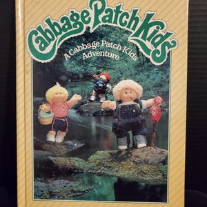 May include: A vintage "Cabbage Patch Kids" photo storybook by Parker Brothers. The cover features the title in green and white, with illustrations of Cabbage Patch Kids dolls in a forest setting. The book has a yellow cover with diagonal lines.