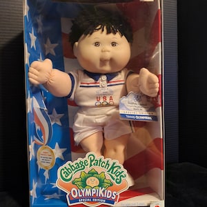May include: A Cabbage Patch Kids OlympiKids Special Edition doll in its original box. The doll is dressed in a white and blue tennis outfit with "USA" printed on the chest. The box features patriotic stars and stripes.