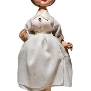 May include: A vintage doll with blonde hair, wearing a white dress and a matching headband. The doll has a painted face with blue eyes and red lips. It stands on a black circular base.