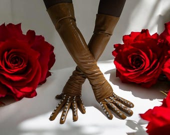 May include: Brown leather opera gloves with rhinestone embellishments on the fingers. The gloves are crossed, with red roses in the background. The gloves are long, extending past the elbows.