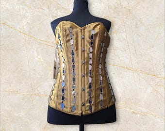 May include: A gold satin corset with a sweetheart neckline and silver mirrored accents. The corset has a busk closure in the front and is decorated with rows of silver mirrored teardrop shapes.
