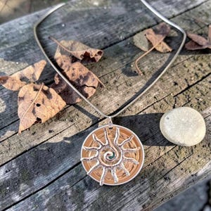 Könnte beinhalten: Eine silberne Halskette mit einem Sonnenanhänger. Der Anhänger hat eine kupferfarbene, kreisförmige Basis mit einem silbernen Sonnenmuster. Die Halskette liegt auf einer Holzoberfläche mit getrockneten Blättern.