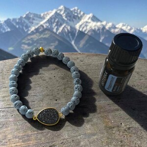 May include: A beaded bracelet featuring light grey porous lava stone beads, a single gold-toned accent bead, and a central teardrop-shaped black druzy charm with a gold-toned rim, positioned next to a small brown bottle labelled dōTERRA Peppermint.