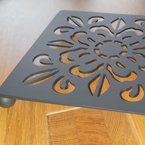Puede incluir: Un salvamanteles cuadrado de metal gris oscuro con un diseño decorativo de corte floral. El salvamanteles tiene una base elevada con cuatro pequeños pies redondeados. El salvamanteles mide aproximadamente 20 cm cuadrados.