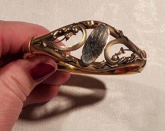 May include: A gold-toned bangle bracelet with an oval-shaped, engraved, silver-toned center piece. The bracelet is decorated with floral designs.