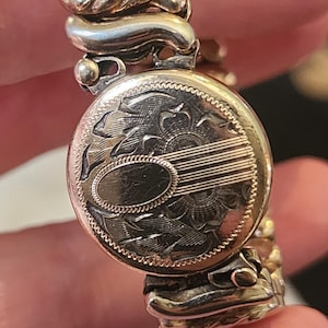 May include: A gold-coloured vintage bracelet with an engraved locket. The locket has a floral design and an oval space. The bracelet has a flexible, expandable design, typical of the era. The metal has a slightly aged appearance.