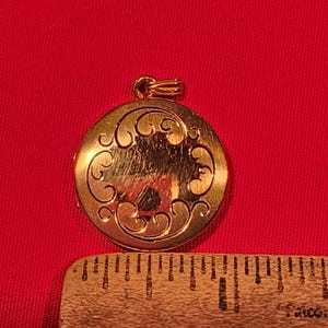 May include: A round gold-toned metal locket pendant featuring an engraved scrollwork pattern on the front surface. The locket is positioned above a wooden ruler, showing the circular pendant has a diameter of approximately 1 inch.