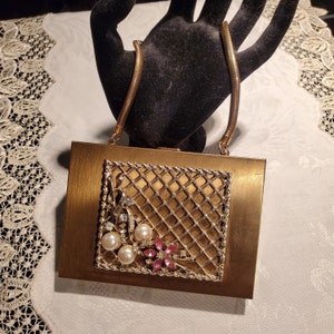 Op de afbeelding: Een gouden metalen clutch met een kettingband. De voorkant van de clutch heeft een decoratief ontwerp van een gouden gaas met roze en witte juwelen.
