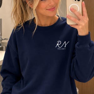 May include: A navy blue crewneck sweatshirt featuring the white embroidered text RN with the name KELSEY printed in a smaller font directly underneath on the upper left chest area. The garment has long sleeves with ribbed cuffs and a ribbed neckline.