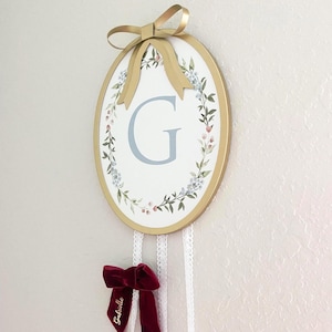 May include: Oval-shaped wall decor with a gold frame and a gold bow at the top. The center features a floral wreath design around a light blue letter "G". A burgundy velvet ribbon with the name "Gabrielle" hangs below.
