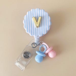 May include: A badge reel with a blue and white striped, scalloped design. A gold letter "V" is centered on the reel. Attached are two pacifier charms, one blue and one pink. A clear plastic clip is also attached.