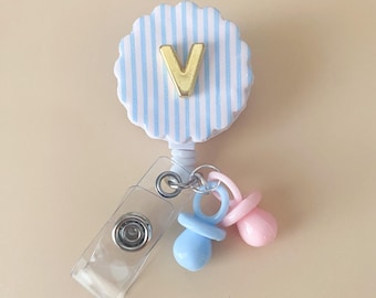 Puede incluir: Un portatarjetas con un diseño festoneado a rayas azules y blancas. La letra dorada "V" está centrada en el portatarjetas. Se adjuntan dos dijes de chupete, uno azul y otro rosa. También se adjunta un clip de plástico transparente.