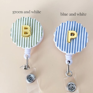 Puede incluir: Dos portatarjetas retr&aacute;ctiles con bordes festoneados. Uno presenta rayas verticales verdes y blancas con una letra B dorada, y el otro rayas verticales azules y blancas con una letra P dorada. Ambos tienen carcasas y clips de pl&aacute;stico blanco.