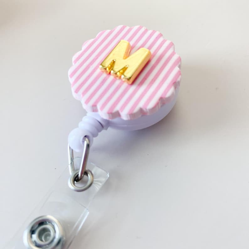 Puede incluir: Un portatarjetas retr&aacute;ctil con una parte superior circular festoneada con un patr&oacute;n de rayas blancas y rosa claro. Una letra M de color dorado brillante est&aacute; centrada en la superficie rayada. El carrete incluye una correa de pl&aacute;stico transparente.