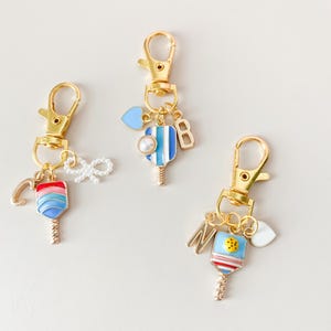 May include: Three gold keychains with colorful paddleball charms. Each charm has a letter, a heart, and a pearl. The letters are C, B, and M.