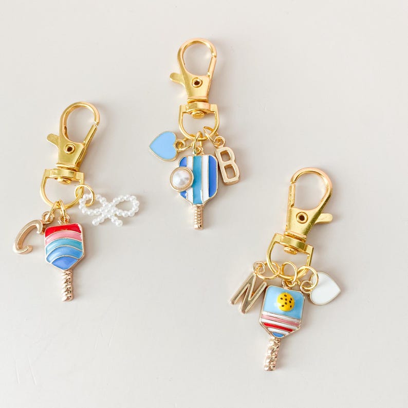 May include: Three gold keychains with colorful paddleball charms. Each charm has a letter, a heart, and a pearl. The letters are C, B, and M.
