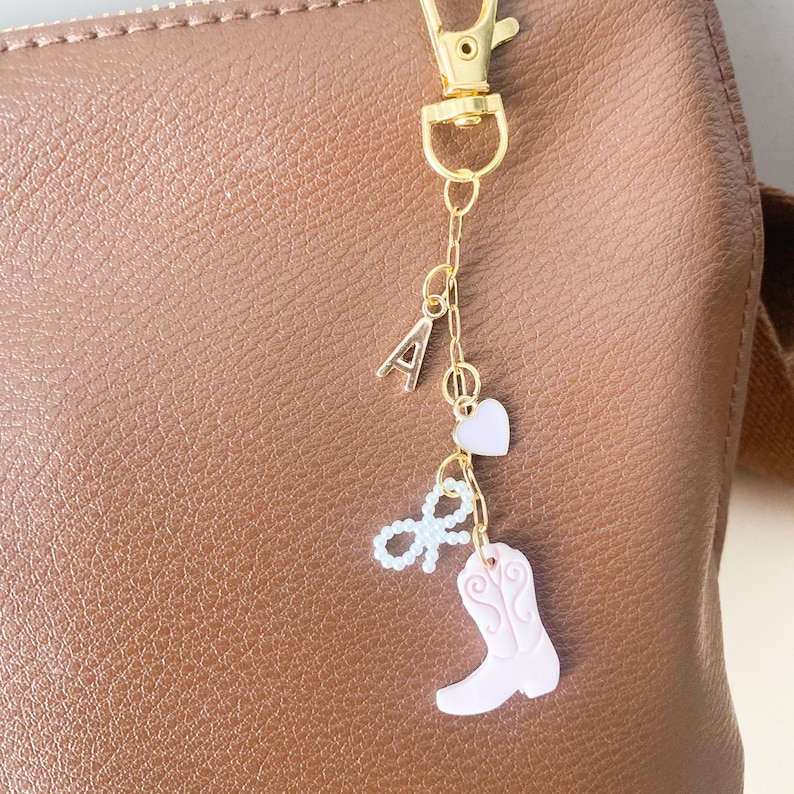 May include: A gold chain keychain with a pink boot charm, a pink heart charm, a white pearl bow charm, and a gold letter A charm.