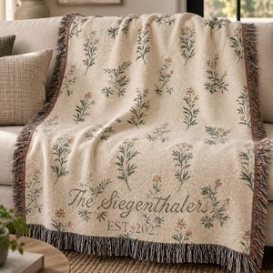 Pode incluir: Uma manta tecida de cor creme com um padr&atilde;o repetitivo de pequenas flores silvestres verdes e cor de laranja. A manta inclui o texto The Siegenthalers e EST. 2027 numa fonte cursiva escura, com franjas decorativas ao longo das extremidades.