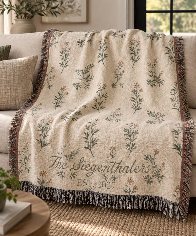 Pode incluir: Uma manta tecida de cor creme com um padr&atilde;o repetitivo de pequenas flores silvestres verdes e cor de laranja. A manta inclui o texto The Siegenthalers e EST. 2027 numa fonte cursiva escura, com franjas decorativas ao longo das extremidades.
