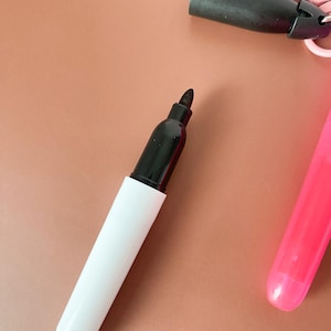 Puede incluir: Un marcador permanente negro con cuerpo blanco y tapa negra. El marcador tiene una punta de fieltro. Cerca, hay una tapa negra separada unida a un clip de pl&aacute;stico rosa, y se ve una parte del cuerpo de un marcador de color rosa brillante.