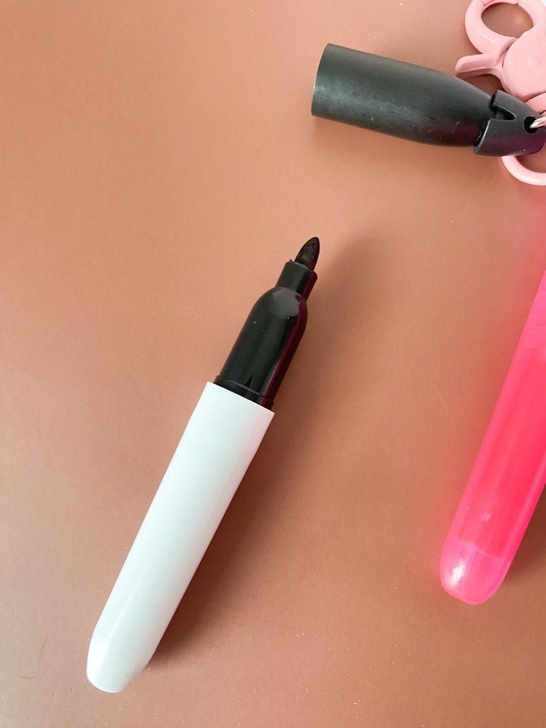 Puede incluir: Un marcador permanente negro con cuerpo blanco y tapa negra. El marcador tiene una punta de fieltro. Cerca, hay una tapa negra separada unida a un clip de pl&aacute;stico rosa, y se ve una parte del cuerpo de un marcador de color rosa brillante.