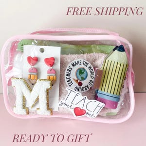 May include: A clear zippered pouch containing a white letter M patch with gold trim, pencil-shaped earrings with red heart tops, a round sticker reading Teachers Make The World Go Around, and a sticker reading Teach your heart out with a red heart.