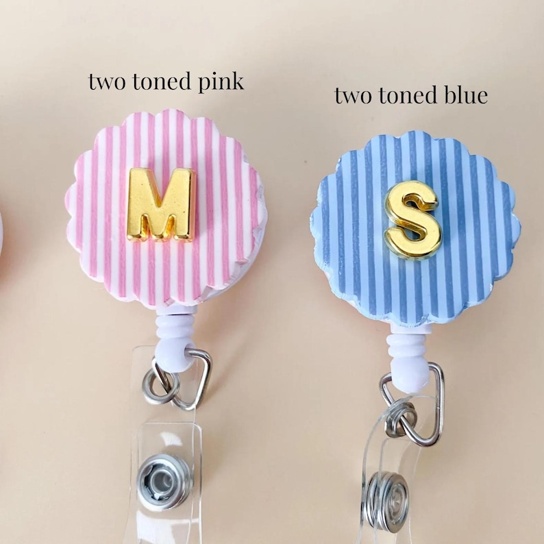 Puede incluir: Dos portatarjetas con bordes festoneados. Uno es de rayas rosas y blancas con una letra M dorada, etiquetado two toned pink. El otro es de rayas azules y blancas con una letra S dorada, etiquetado two toned blue. Ambos tienen correas y clips.