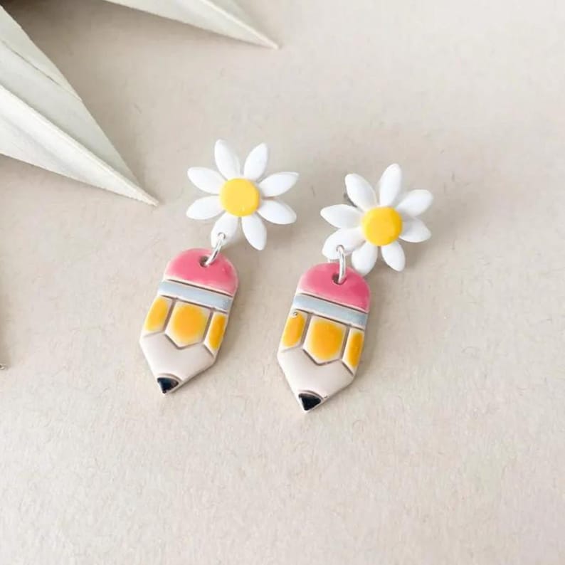 May include: A pair of dangle earrings featuring a white daisy stud with a yellow center, connected to a pencil-shaped charm. The pencil charms are pink, yellow, and white with a black tip. The earrings are on a neutral background.