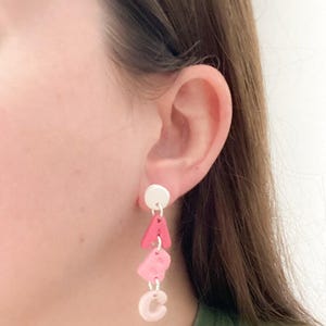 May include: A dangle earring with a white stud and three pink letter charms spelling "ABC". The letters are in varying shades of pink, with the letter "A" being a darker shade than the other letters.